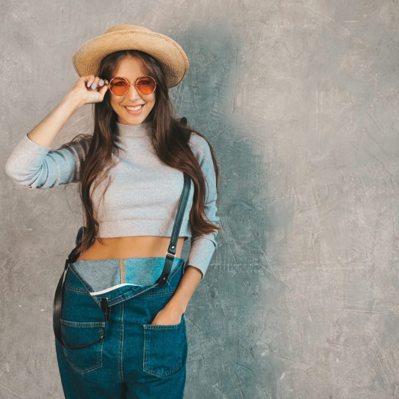 Young beautiful smiling woman looking at camera. Trendy girl in casual summer overalls clothes and hat. Funny and positive female posing near gray wall in studio